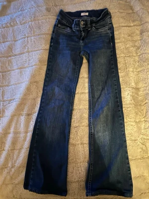 Blå bootcut jeans från Nelly - Snygga blå jeans från Nelly med bootcut passform och låg midja. Jeansen har klassisk femficksdesign, dubbla silverknappar i midjan och coola broderade detaljer på bakfickorna. Materialet är jeans med en skön stretch och slitningar för en avslappnad look. Upplagda till en 155 cm lång tjej.