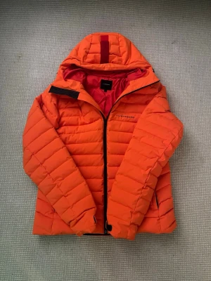 Orange pufferjacka från J.Lindeberg - Säljer en fet orange pufferjacka från J.Lindeberg i storlek M. Jackan har huva, svarta detaljer och dragkedja framtill. Den är quiltad och har ett cleant, sportigt vibe med logga på bröstet. Perfekt för kalla dagar när du vill sticka ut.