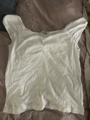 Vit ribbad topp från AWARE - Säljer en vit topp från AWARE i storlek M. Toppen har breda axelband, v-ringad hals och ett ribbat mönster med diskreta vertikala ränder. Materialet känns mjukt och luftigt, perfekt för varmare dagar.💗