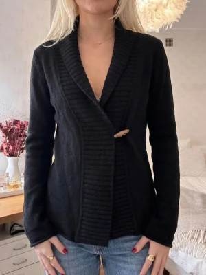 Svart kofta från Boomerang, storlek L - Säljer en svart kofta från Boomerang i storlek L men mer som en S/M. Koftan har en bred ribbad krage och stängs med en snygg träknapp framtill. Materialet är mjukt och känns riktigt skönt mot huden. Perfekt att slänga på sig när det är lite kyligt ute.