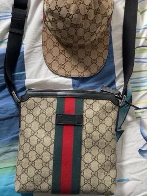Gucci axelväska med GG-mönster äkta  - Säljer en snygg axelväska från Gucci med klassiskt GG-mönster i beige och brunt. Väskan har bred axelrem, detaljer i svart skinn och den ikoniska gröna och röda randen i mitten. Perfekt för dig som vill ha en stilren och lyxig accessoar.