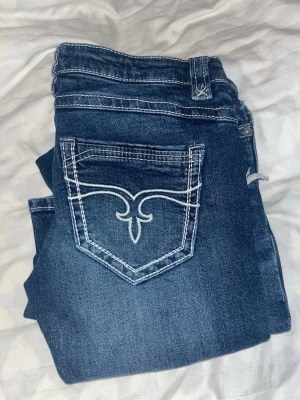 Blå bootcut jeans från Gina Tricot - Snygga blå jeans från Gina Tricot i bootcut-modell med slitna detaljer och kontrastsydda sömmar. Jeansen har en klassisk femficksdesign och dekorativ broderad bakficka. Perfekt för dig som gillar en avslappnad och trendig look.