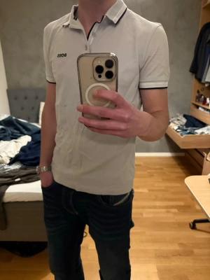 Vit piké från BOSS med svarta detaljer - Säljer en stilren pikétröja från BOSS med svarta tunna ränder på krage och ärmslut. Tröjan har korta ärmar, klassisk krage och knappslå framtill. Materialet är mjuk bomull och loggan är broderad på bröstet. Perfekt för dig som gillar enkel och snygg design.