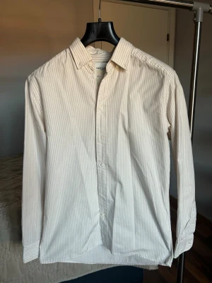 Beige randig skjorta från These Glory Days - Snygg beige skjorta med tunna vita ränder från These Glory Days. Klassisk krage, långärmad och knäppning framtill. Skjortan är gjord i mjuk bomull och har en relaxed passform som är perfekt för en chill stil.