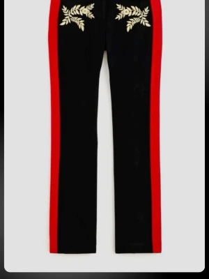 Rabanne x H&M svarta broderade byxor - Svarta kostymbyxor med breda röda sidor, detaljerade med guldiga broderade blommor och blad framtill. Byxorna har guldfärgade knappar med emblem och är tillverkade i ett ulligt material. Snygg rak passform och exklusiv design.