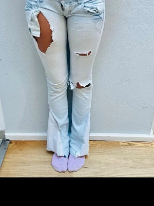 Ljusa bootcut jeans med slitningar - Säljer ett par ljusa jeans med bootcut-snitt och råa slitningar på låren och knäna. Byxorna har klassisk femficksdesign, väldigt låg  midja och är tillverkade i jeansmaterial. Perfekt för dig som gillar en edgy och avslappnad stil.
