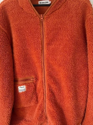 Orange teddyjacka från Resteröds - Mysig orange teddyjacka från Resteröds med hel dragkedja framtill och en liten ficka med dragkedja på sidan. Jackan har långärmade ärmar och är tillverkad i fluffigt fleece-material som ger en skön och avslappnad vibe.
