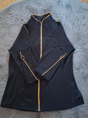 Svart träningsjacka från Bodyzone - Svart träningsjacka med gul kontrastdragkedja och detaljer längs ärmarna. Jackan har hög krage och två sidofickor med dragkedja. Materialet är mjukt och stretchigt, perfekt för aktiva dagar. BZAW-logga i gult på ryggen. Snygg och sportig look.