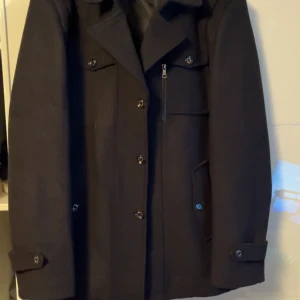 Svart rock  - Säljer en svart rock med klassisk krage, flera fickor framtill och snygga knappar längs hela framsidan. Jackan har lång ärm med knappdetaljer vid ärmslut och är tillverkad i ett tjockare ullmaterial. Perfekt för kyliga dagar och stilren look.