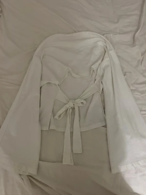 Öppen rygg tröja  - Säljer en vit omlottopp från Nelly i storlek S. Toppen har långa ärmar, rund halsringning och snygg knytning i ryggen som ger en öppen detalj. Materialet känns mjukt och lätt, perfekt för lager på lager eller som statement-piece.