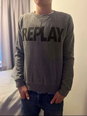 Grå sweatshirt från Replay - Säljer en grå sweatshirt från Replay med stor svart logotyp på bröstet. Tröjan har rund hals, långa ärmar och ribbade muddar vid ärmslut och nederkant. Perfekt för chill dagar och enkel att matcha med jeans eller joggers.