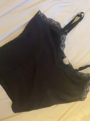 Svart linnetopp med spets från Divided  - Säljer en svart linnetopp från Divided i storlek XS. Toppen har tunna axelband och en snygg spetskant längs halsringningen som ger en extra touch.