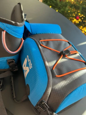 Blå och svart Non-stop Dogwear midjebälte + väska - Säljer en sportig och funktionellt midjebälte från Non-stop Dogwear i storlek L. Den är blå och svart med orange detaljer och har flera fästen, spännen och elastiska snören framtill. Materialet är slitstark syntet och polyester, perfekt för aktiva hundpromenader eller träning.