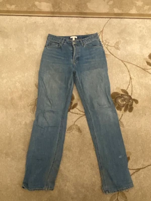 Blå raka jeans från H&M, strl 40 - Säljer ett par klassiska blå jeans från H&M i storlek 40. Jeansen har rak passform, hög midja och stängs med flera silverfärgade knappar framtill. Materialet är jeans och de har fem fickor. Perfekt för dig som gillar enkel och tidlös stil.