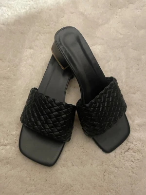 Svarta flätade sandaler med låg klack - Säljer ett par svarta sandaler med bred, flätad rem i skinnimitation och fyrkantig tå. Skorna har låg klack och öppen design, perfekta för sommarens alla tillfällen. Stilrena och trendiga med en chunky känsla.