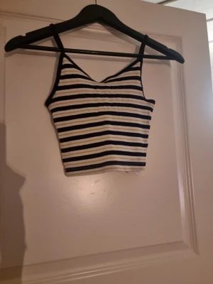 Randig ribbad linnetopp - Säljer en croppad linnetopp med smala axelband och ribbad struktur. Toppen har svarta, vita och ljusgula horisontella ränder och är i ett mjukt stretchigt material. Perfekt till sommaren och enkel att matcha med jeans eller kjol.