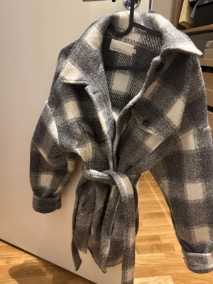 Rutig jacka från NLY Trend - Säljer en rutig jacka från NLY Trend i grått och vitt. Jackan har knappar framtill, bröstfickor och ett bälte i midjan för extra stilpoäng. Perfekt för lager-på-lager och chill vibe. Materialet är mjukt och känns ulligt. Använd många gånger och är nopprig, se bild.