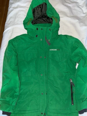 Grön jacka från Didriksons - Säljer en grön jacka från Didriksons med huva och flera fickor framtill. Jackan har dragkedja och tryckknappar, samt justerbar huva. Perfekt för kalla dagar och har en clean, sportig look.