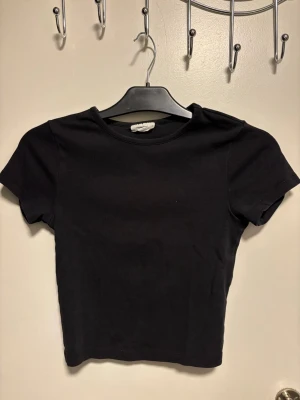 Svart croppad t-shirt - En enkel svart croppad t-shirt med rund hals och korta ärmar. Toppen är gjord i mjuk bomull och har en stilren, minimalistisk look som är lätt att matcha med olika outfits. Perfekt för dig som gillar basic plagg med modern passform.