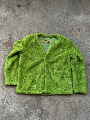 Grön teddyjacka från CPFM.XYZ - Säljer en unik grön teddyjacka från CPFM.XYZ med fluffig textur och två fickor framtill. Jackan har en avslappnad passform och stängs med knappar. Perfekt för dig som vill sticka ut med en färgglad och mysig look.