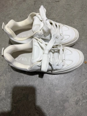 Vita sneakers från Zara strl 43 - Säljer ett par fräscha vita sneakers från Zara i storlek 43. Skorna har rund tå, platt sula och bred snörning. Ovandelen är i syntetmaterial och textil, med en stilren och enkel design som passar till många olika outfits.