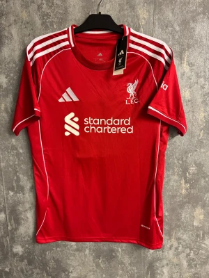 Liverpool tröja M. Salah #11 Adidas - Röd Liverpool FC matchtröja från Adidas med vita detaljer och tryck. Framsidan har klubbmärke och sponsorlogga, baksidan har M. Salah och nummer 11 i vitt. Tröjan har korta ärmar och klassisk rund hals. Perfekt för fotbollsfans som vill visa sitt stöd.