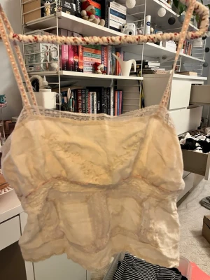 Vit spetsdetaljerad linnetopp - Säljer en söt vit linnetopp med tunna axelband och spetsdetaljer längs kanterna. Toppen har en lätt volang nedtill och dekorativa sömmar med ljusrosa inslag. Perfekt för varma dagar och sommarhäng. Saknar en liten rosett på framsidan