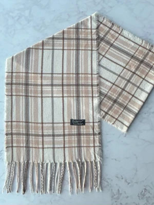 Beige rutig Burberrys väst i lammull - Säljer en klassisk väst från Burberrys i mjuk 100% lammull. Västen har ett rutigt mönster i beige, brunt och grått med fransar längs nederkanten. Perfekt för dig som gillar tidlös design och vill ha något som sticker ut lite extra.