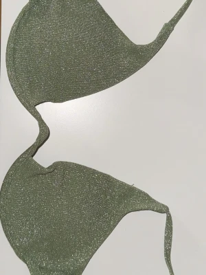 Glittrig grön bikini topp - Snygg grön bikini topp med glittrigt tyg och smala axelband. Bikini toppen har en triangelformad design och knytning i ryggen för justerbar passform. Perfekt för dig som gillar att sticka ut med lite extra glitter i outfiten. Den har byglar och stel form.