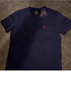 Marinblå t-shirt från Polo Ralph Lauren - Snygg marinblå t-shirt från Polo Ralph Lauren med klassisk röd logga broderad på bröstet. T-shirten har korta ärmar och är gjord i mjuk bomull som känns skön mot huden. Perfekt för dig som gillar stilrena och tidlösa plagg.