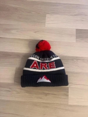 Blå Åre mössa med tofs och brodyr - Säljer en blå stickad Åre-mössa med vit och röd text, stor tofs upptill och broderat bergmotiv framtill. Mössan har uppvikt kant och texten 'SWEDEN' samt 'ÅRE' i rött. Perfekt för kalla dagar och vintersport.
