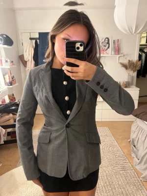 Grå kavaj med klassisk passform - Snygg grå kavaj med klassiska fickor och enkla svarta knappar. Passar till både jeans och kjol. Storlek s men skulle säga att den är mer som en XS