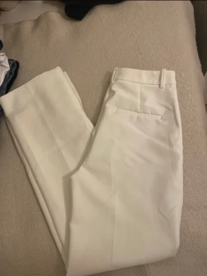 Vita kostymbyxor från Zara - Snygga vita kostymbyxor från Zara med hög midja och raka ben. Byxorna har klassiska bälteshällor och pressveck framtill för en clean look. Materialet är mjukt och känns lyxigt, perfekt för dig som gillar stilrena outfits.