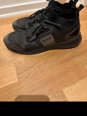 Svarta sneakers från BOSS - Säljer ett par svarta sneakers från BOSS med stickad strumpkrage och snörning. Skorna har en mix av syntet och stickat material, samt en platt sula och rund tå. Snygg detalj med BOSS-logga på sidan och diskreta gröna inslag vid hälen.