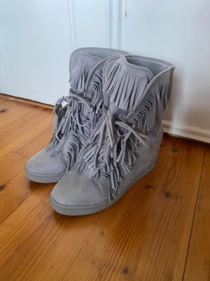 Super snygga franskor - Säljer ett par grå boots med kilklack och rund tå. Skorna har snygga fransdetaljer över hela ovandelen och snörning framtill. Materialet är mocka och de har en platt sula med kilklack för extra höjd. Perfekt för dig som gillar statement-detaljer.