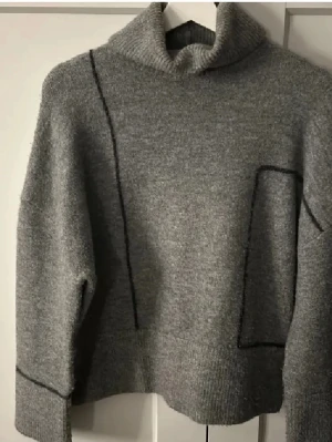 Grå Zara tröja  - Säljer en grå polotröja med svarta linjer som ger en cool och modern look. Tröjan har en hög krage och är långärmad med ribbade muddar. Materialet är stickat och känns mjukt mot huden. Perfekt för dig som gillar stilrena och enkla plagg.