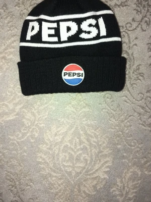 Svart stickad Pepsi mössa - Säljer en svart stickad mössa med vit Pepsi-logga och ett broderat Pepsi-märke framtill. Mössan har bred uppvikt kant och är riktigt snygg för dig som gillar retro och streetwear. Perfekt för kalla dagar och enkel att matcha med din outfit. Limited Edition pepsi mössa.