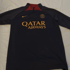PSG långärmad träningströja Nike S - Paris Saint-Germain långärmad träningströja från Nike i mörkblått med röda detaljer vid kragen. Guldiga tryck med klubbmärke, Nike-logga och Qatar Airways på bröstet. Tillverkad i Dri-FIT-material som andas och har halv dragkedja framtill. Nytt skick 9,9/10!🔥