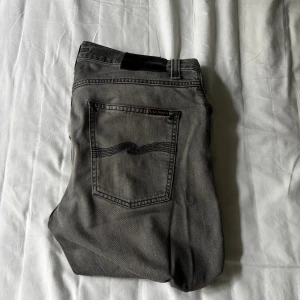 Grå jeans från Nudie Jeans, 31/34 - Snygga grå jeans från Nudie Jeans med klassisk femficksdesign och svarta broderade vågmönster på bakfickorna. Jeansen har en smal passform och är tillverkade i mjukt bomullsmaterial. Perfekt för dig som gillar stilrena och tidlösa jeans.