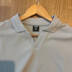 Pike - Snygg ljusgrå piké t-shirt från FSBN med klassisk krage och v-ringning. T-shirten har korta ärmar och är tillverkad i ett mjukt material som känns skönt mot huden. Perfekt för en clean och stilren look.