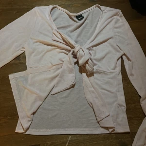 Ljusrosa knytblus från Gina Tricot - Supersöt ljusrosa blus från Gina Tricot med långa ärmar och knyt framtill. Tunn och mjuk i materialet, perfekt för lager-på-lager eller som statement-piece. Snygg v-ringning och lätt transparent tyg ger en trendig vibe.