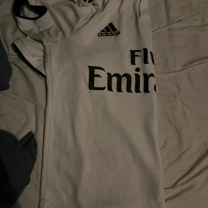 Vit Real Madrid fotbollströja Adidas - Säljer en vit Real Madrid fotbollströja från Adidas med svart tryck och klassiska svarta ränder på axeln. Tröjan har korta ärmar och är tillverkad i ett lätt och ventilerande material, perfekt för träning eller match. Fly Emirates-logga på bröstet.