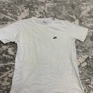 Vit t-shirt från Sport Master M - Vit t-shirt från Sport Master i klassisk modell med rund hals och korta ärmar. Liten Nike-logga broderad på bröstet i mörkblått. Tillverkad i mjuk bomull, perfekt för en clean och enkel stil.