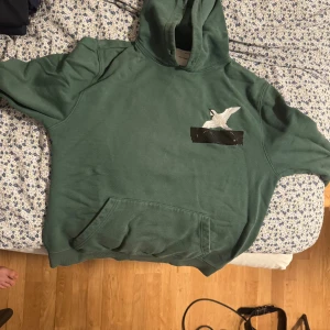 Arigati-Grön Tape Hoodie. Storlek M. Saknar snöre - Säljer en grön hoodie med huva och magficka. På bröstet finns en broderad vit fågel ovanför ett svart band. Hoodien har en avslappnad passform och är perfekt för chill dagar. Materialet känns mjukt och skönt mot huden.