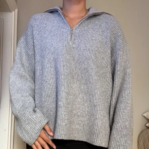 Grå ribbstickad half zip-tröja - Mysig grå ribbstickad tröja med half zip-dragkedja och krage. Tröjan har en loose passform och långa ärmar, perfekt för lager-på-lager. 