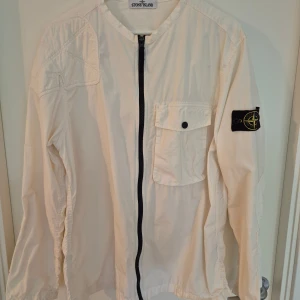 Vit overshirt från Stone Island - Säljer en stilren vit overshirt från Stone Island med klassisk patch på ärmen. Jackan har en rak passform, bröstficka med knapp och hel dragkedja framtill. Tillverkad i bomull och perfekt för lager-på-lager. Enkelt och cleant plagg som lyfter din outfit.