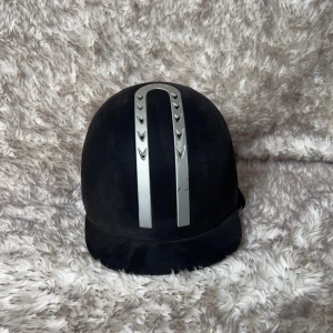 Svart ridhjälm från HS equestrian med silverdetaljer - Snygg svart ridhjälm från SH med två breda silverfärgade ränder. Mjuk insida för extra komfort. Om man zoomar in på bild 1 så ser man att det det finns ett litet hack på den vänstra sidan av skärmen samt lite skrapmärken på silverränderna.