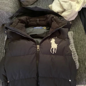 Svart dunväst från Polo Ralph Lauren - Svart dunväst från Polo Ralph Lauren med stor broderad logga på bröstet. Västen har hög krage, dragkedja framtill och två fickor med dragkedja. Klassisk och stilren modell som är perfekt för lager-på-lager under kyliga dagar.