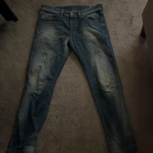 Diesel Buster jeans blå W32 - Säljer ett par blå Diesel Buster jeans med snyggt slitna detaljer och klassisk femficksdesign. Jeansen har regular slim-tapered passform och är tillverkade i stretchigt denim för extra komfort. Perfekta för dig som gillar en modern men avslappnad look.