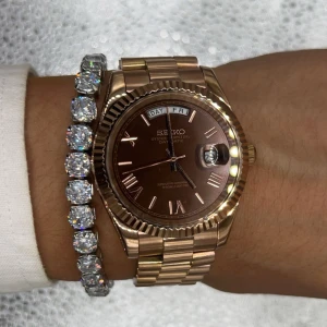Seiko Day-Date guldbrun klocka - Säljer en stilren Seiko Oyster Perpetual Day-Date med guldigt armband och boett. Urtavlan är brun med romerska siffror och dag- och datumvisning. Klockan har en klassisk rund form och länkarmband i metall som ger en lyxig vibe.